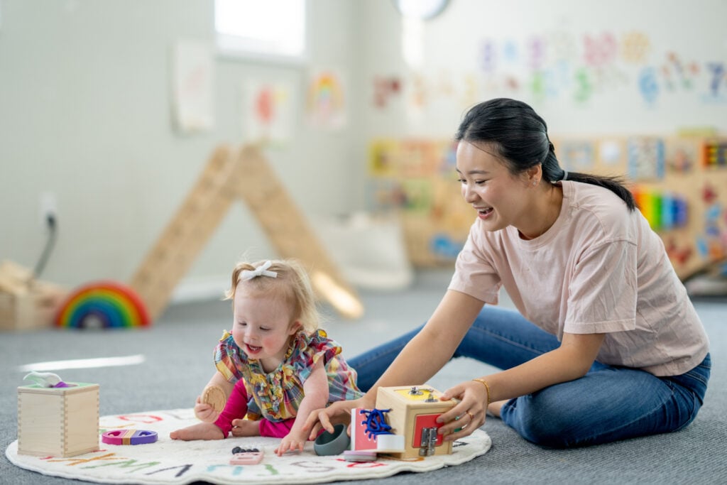 A sweet little girl sits on the floor with her childcare provider as she plays with a variety of toys. She is dressed casually and has a neutral expression on her face as the two interact together.