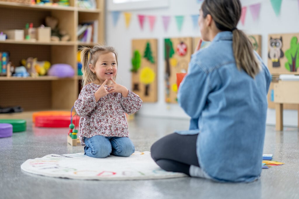 A female Speech therapist sits with a little girl on the floor as she works with her on phonetics. The therapist is making a sound and holding her finger to her mouth for the little girl to mimic.
